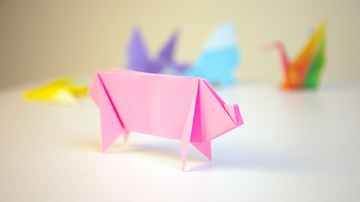 In the foreground is a pink origami pig shape. In the background, out of focus, are various multi-coloured origami animal shapes