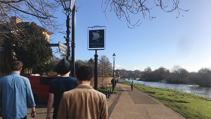 Image of the backs of three men walking along Richmond's riverside towards a pub sign. It is a bright sunny day.