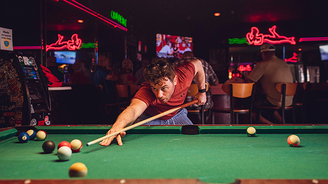 Men’s Club – Cue the Competition. Man playing pool in dimley lit sports-style bar.