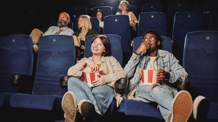 A couple sit, legs up at a cinema, smiling and eating popcorn