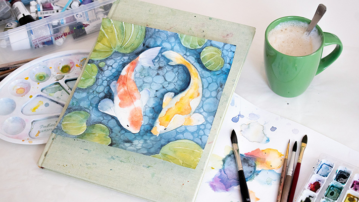 A hand painted image of two fish. A mug of coffee and paint materials surround the painting