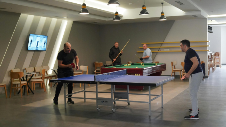 In the foreground, men play table tennis together. In the background, two men playing pool